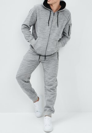 Man draagt lichtgrijze, gemêleerde zip-up hoodie en bijpassende joggingbroek met witte sneakers, leunend tegen een witte muur.