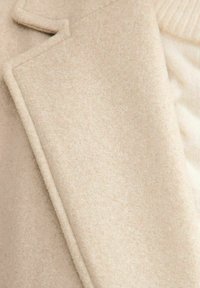 Abrigo de lana beige con un acabado texturizado, que presenta líneas limpias y un cuello marcado, parcialmente superpuesto a un suave suéter de punto color crema.