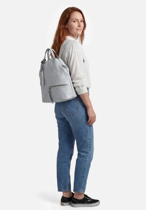 Jeune femme aux longs cheveux roux portant une chemise blanche, un jean bleu, des baskets noires, avec un sac à dos en cuir gris clair porté sur une épaule.