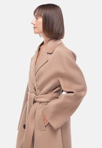 Cappotto di lana beige con vita stretta, ampi rever e maniche lunghe. Presenta bottoni e una texture morbida. Mostrato in profilo laterale.