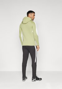 Hanorac cu glugă de culoare verde deschis, cu mâneci albe, purtat alături de o pereche de pantaloni joggeri negri cu dungi albe pe laterale și pantofi sport negri.