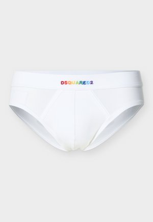 Junger Mann steht barfuß, trägt weiße Slips mit dem "DSQUARED2"-Logo auf dem Bund vor einfachem, hellem Hintergrund.