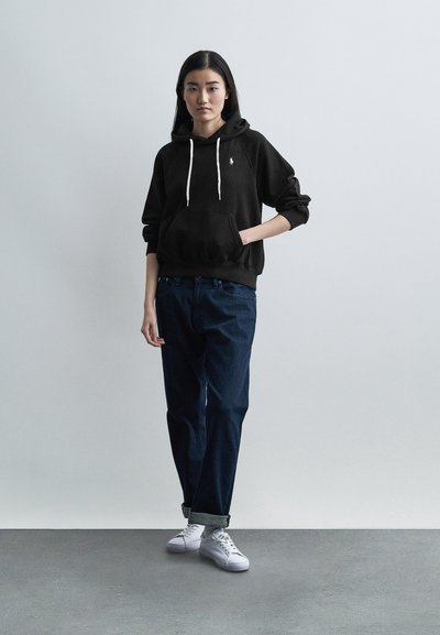 Γυναίκα που φοράει μαύρη κουκούλα, σκούρο μπλε τζιν με γυρισμένα μπατζάκια και λευκά αθλητικά παπούτσια, στέκεται μπροστά σε απλό ανοιχτό γκρι φόντο.