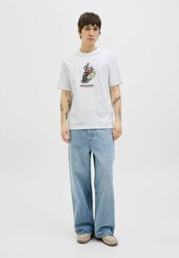 Tricou alb din bumbac, cu un print grafic al unui motociclist și text, asociat cu blugi din denim cu picioare largi de culoare albastru deschis.