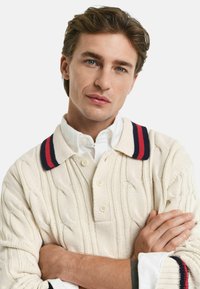 Creme kabelstrik sweater med kravehals, der har marineblå og røde striber. Inkluderer knapper og en kontrasterende hvid skjorte under.