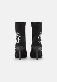 Czarne botki za kostkę na wysokim obcasie typu stiletto, wykonane z elastycznego materiału. Posiadają zamek błyskawiczny i wyraźny biały napis po zewnętrznej stronie.