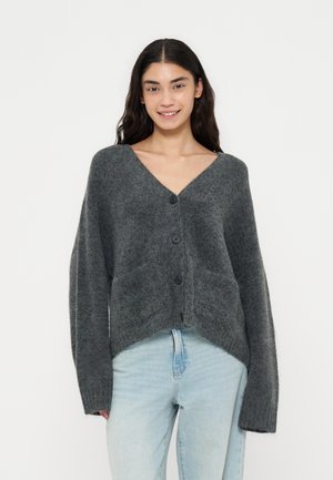 Kvinde med langt mørkt hår iført en løs mørkegrå cardigan med knapper og lyseblå jeans, stående mod en ensfarvet hvid baggrund.