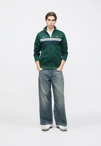 Νέος άνδρας στέκεται με τα χέρια στις τσέπες, φορώντας πράσινο αθλητικό μπουφάν με φερμουάρ και λευκές ρίγες, φαρδιά ξεβάμματα τζιν και άσπρα αθλητικά παπούτσια.