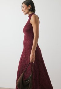 Bordeaux spetsklänning med hög halsringning och utsvängd kjol. Har intrikata blommönster och en slät textur, som matchas med en brun väska.