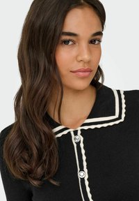 Crna rebrasta pletena kardigan s oštrim, kontrastnim bijelim ovratnikom i zatvaranjem na dugmad. Ima okrugla dugmad i prilagođeni kroj.