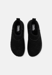 Fekete slip-on cipők, texturált, bolyhos felső résszel és kerek orral. Jellemzője a kontrasztos sima fekete talp és egy kis húzófül a saroknál.