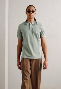 Lys grønn bomullspolo med krage og brodert logo, kombinert med løse beige bukser, båret av en person med fletter.