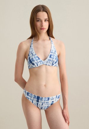Frau mit langen braunen Haaren, die einen blau-weißen Batik-Halterneck-Bikini vor einem schlichten beigen Hintergrund trägt.