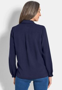 Blusa a maniche lunghe blu navy con piega posteriore, colletto e polsini con bottoni, realizzata in tessuto liscio. Abbinata a jeans aderenti.