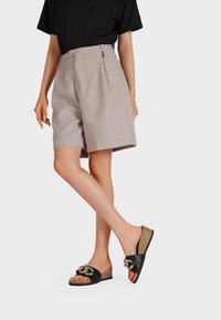 Infradito neri e dorati con una texture intrecciata, dotate di un accento decorativo a catena; indossati con pantaloni corti grigi chiari su misura e una maglietta nera.