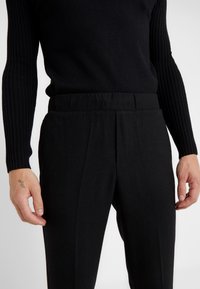 Homme portant un pull côtelé noir à manches longues et un pantalon noir taille haute, les mains détendues le long du corps, montrant un petit tatouage à la main gauche.