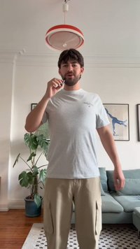 Un hombre con una camiseta gris claro y pantalones de cargo beige sostiene un dispositivo negro. Hay una planta verde y un sofá azul en el fondo.