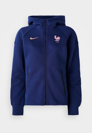 Haut de survêtement zippé bleu marine en tissu doux, avec une poche kangourou, un blason de football français et un logo Nike en corail.
