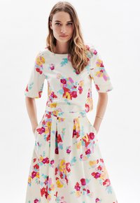 Ensemble floral en deux pièces comprenant un top crème à manches courtes et une jupe assortie. Les deux pièces présentent des motifs de fleurs multicolores et des plis.