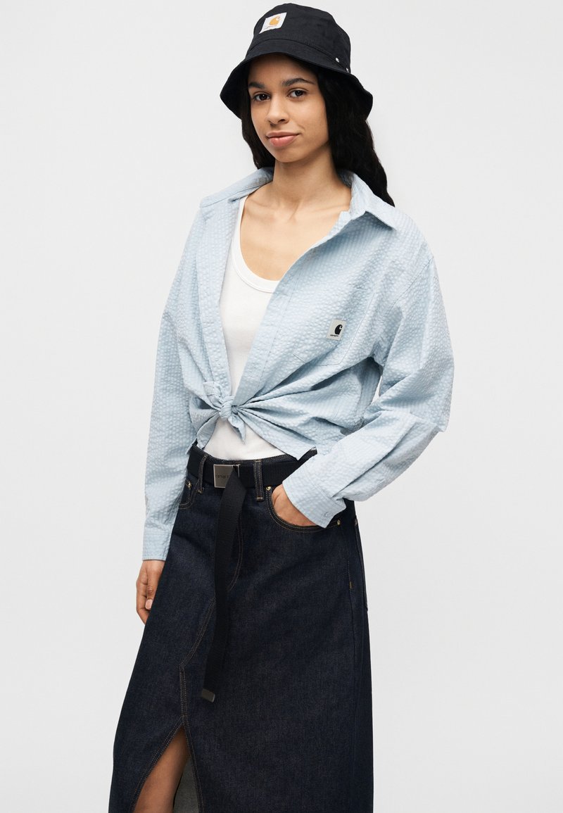 Chemise à manches longues bleue texturée boutonnée, nouée à la taille, associée à une jupe en denim foncé avec une fente avant et une ceinture noire. Accent d'un chapeau seau noir.
