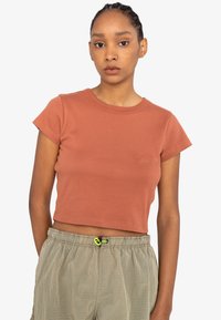 Camiseta cropped de color óxido con mangas cortas y cuello redondo. Fabricada en material acanalado, presenta un discreto bordado del logo en el pecho.