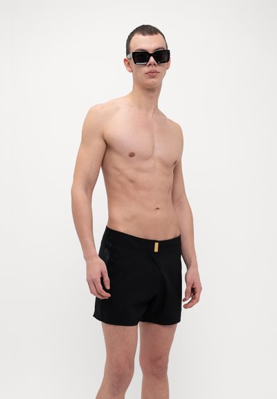 Bañador negro con un acento dorado en la cintura, con un diseño elegante y bolsillos laterales. Modelo con gafas de sol negras, sin camisa.