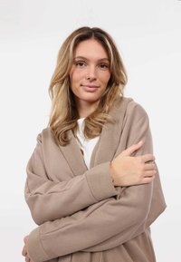 Accappatoio in pile beige con colletto e polsini strutturati. Presenta una silhouette accogliente, maniche lunghe e un design avvolgente. Nessun motivo o dettagli aggiuntivi.