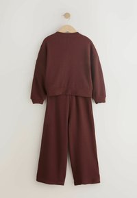 Dwuczęściowy bordowy strój, składający się z teksturowanego dzianinowego topu z opuszczonymi ramionami oraz pasujących szerokich spodni. Zawieszony na drewnianym haczyku.