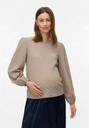 Beige textuur blouse met opgeblazen lange mouwen, ronde halslijn en gathered zoom. Gecombineerd met een donkere rok, staat het model met een hand op de buik.