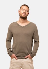 V-Ausschnitt, langärmliger Pullover aus einem weichen, strukturierten beigen Stoff. Gerippte Bündchen und Saum, lockere Passform. Über einer hellen Hose getragen.