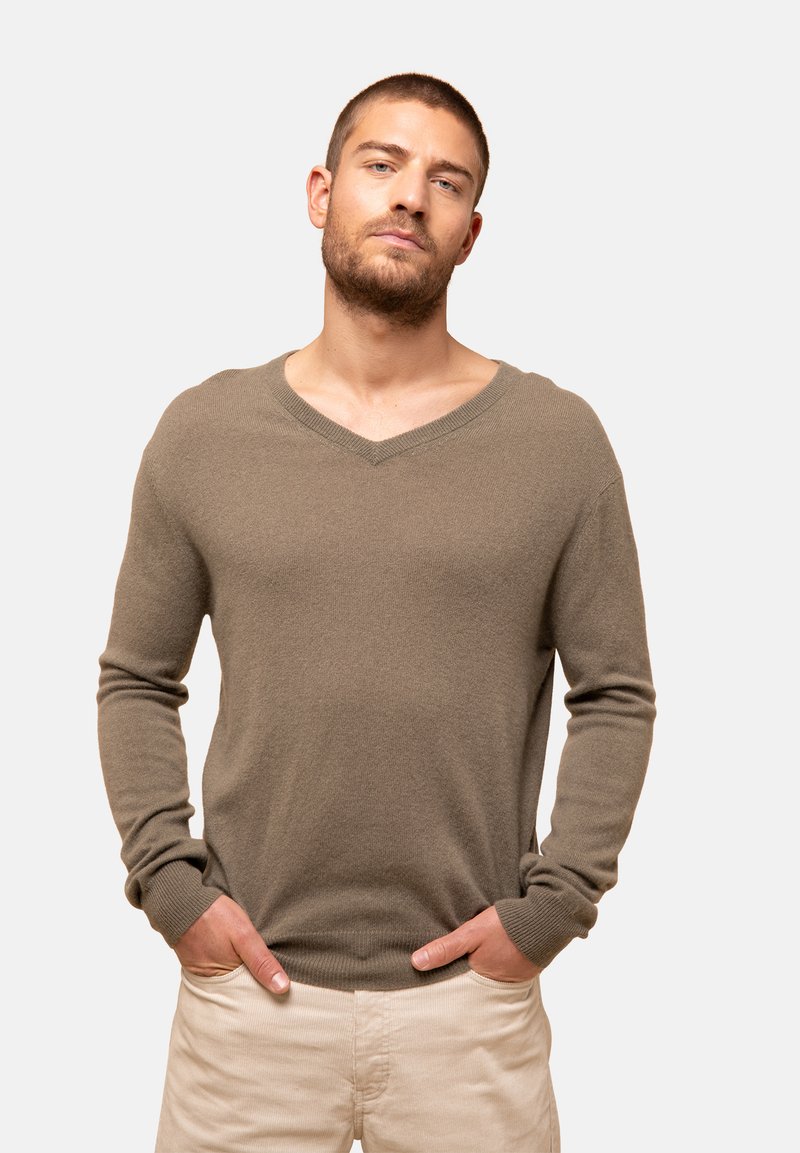 V-Ausschnitt, langärmliger Pullover aus einem weichen, strukturierten beigen Stoff. Gerippte Bündchen und Saum, lockere Passform. Über einer hellen Hose getragen.