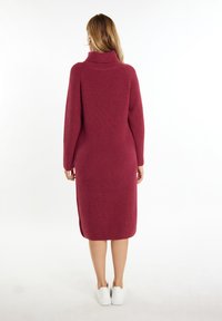 Burgunderfarbenes Strickkleid mit hohem Kragen, langen Ärmeln und seitlichen Schlitzen. Getragen mit weißen Sneakers, die ein strukturiertes, geripptes Design aufweisen.