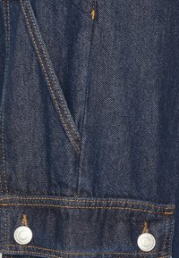 Jeansoverall i mörkblått tyg, med kontrasterande orange sömmar och silverfärgad metalluttagsdetalj med tryckknappar vid fållen.