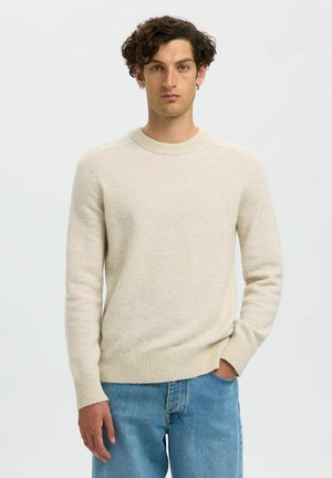 Suéter de punto beige hecho de lana suave, con un escote redondo y puños acanalados. Combinado con jeans de mezclilla azul claro.