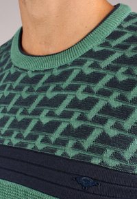 Sweter dzianinowy z misternym wzorem w odcieniach zieleni i granatu, z okrągłym dekoltem, żebrowanymi mankietami oraz małym logo przy dolnym brzegu.