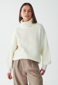 Maglione bianco a collo alto lavorato a maglia con un motivo testurizzato e maniche scese, abbinato a pantaloni a vita alta marrone chiaro.