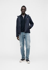 Ung mand med fletninger iført en marineblå jakke, sort sweatshirts med lynlås, lyse revnede jeans og sorte sneakers, stående mod en hvid baggrund.