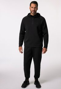 Svart hoodie med halvdragkedja, tillsammans med matchande joggers. Slätt, mjukt tyg med ribbade ärmslut och sidofickor. Svarta sportskor.