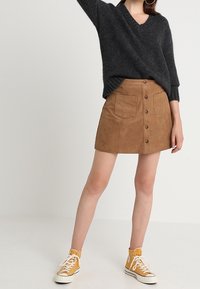Brauner Wildleder-Minirock mit vorderem Knopfverschluss und zwei Taschen, kombiniert mit einem dunkelgrauen Strickpullover und gelben High-Top-Sneakers.