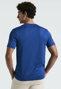 T-shirt bleu royal à manches courtes en tissu lisse et léger ; présente un col rond et une emmanchure arrondie à l'arrière.