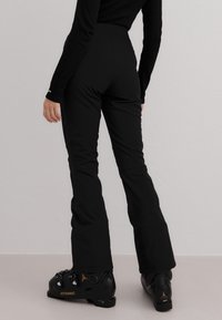 Person iført svarte, tettsittende skibukser og svarte Atomic-skistøvler står mot en enkel lys bakgrunn.