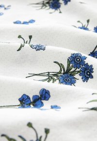 Weißer Stoff mit blauer floralem Stickerei, mit verschiedenen Blumenformen und grünen Stielen, vor einem subtilen Pünktchen-Hintergrund.