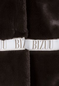 Donkerbruin nepbont stof met een zachte textuur. Een witte lint met "BIZUU" in beige letters, wat een contrasterend accent biedt.