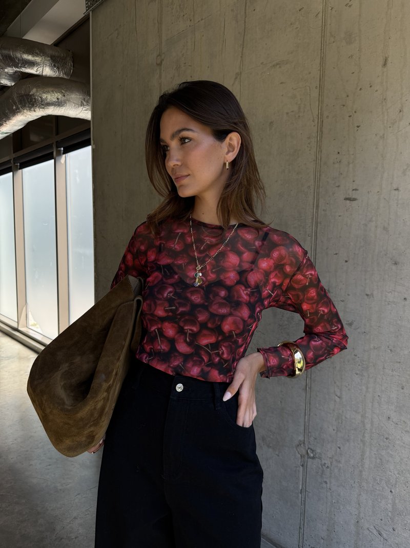 Langärmliges Oberteil mit Kirschmuster, schwarze hochtaillierte Hose und braune Wildlederhandtasche. Goldarmband und Ohrringe sichtbar.