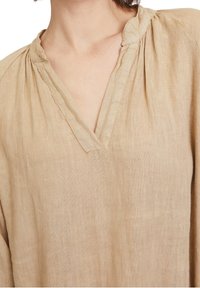 Beige linned tunika, jossa on V-pääntie, ristikohtaiset yksityiskohdat olkapäillä ja rento istuvuus. Kankaan rakenne vaikuttaa pehmeältä ja kevyeltä.