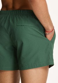 Zöld fürdőnadrág texturált mintával, rugalmas derékpánttal és oldalsó zsebekkel, enyhe fényességgel.