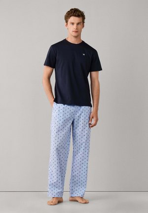 Man staat op blote voeten, draagt een zwart T-shirt met korte mouwen en lichtblauwe pyjamabroek met kleine donkere patronen tegen een effen achtergrond.