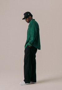 Camicia verde a maniche lunghe, textured, con dettagli in bottoni, abbinata a pantaloni neri a gamba larga e scarpe bianche casual. Un cappellino nero completa il look.