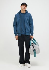 Plava zip-zatvarača preko odgovarajuće majice, uparen sa tamnim hlačama; drži sivu torbu s teget trakama. Jednostavne tenisice upotpunjuju izgled.