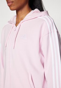 Růžová mikina na zip s bílými pruhy na ramenou, nastavitelnou kapucí a předními kapsami. Materiál vypadá měkký a lehký. Logo Adidas na prsou.